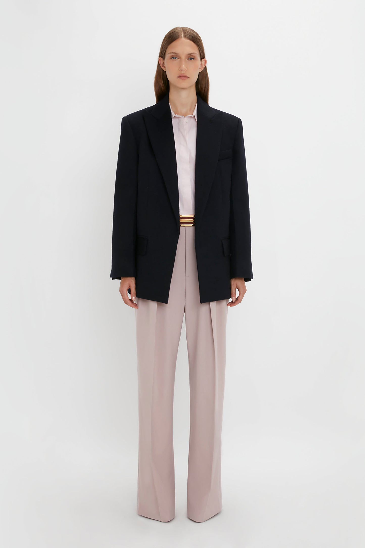 A person stands against a plain white background wearing a Peak Lapel Jacket In Midnight over a light-colored shirt with light pink pants, evoking a contemporary feel. They face forward with a neutral expression, capturing the essence of Victoria Beckham's sophisticated style.