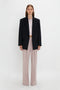 A person stands against a plain white background wearing a Peak Lapel Jacket In Midnight over a light-colored shirt with light pink pants, evoking a contemporary feel. They face forward with a neutral expression, capturing the essence of Victoria Beckham's sophisticated style.