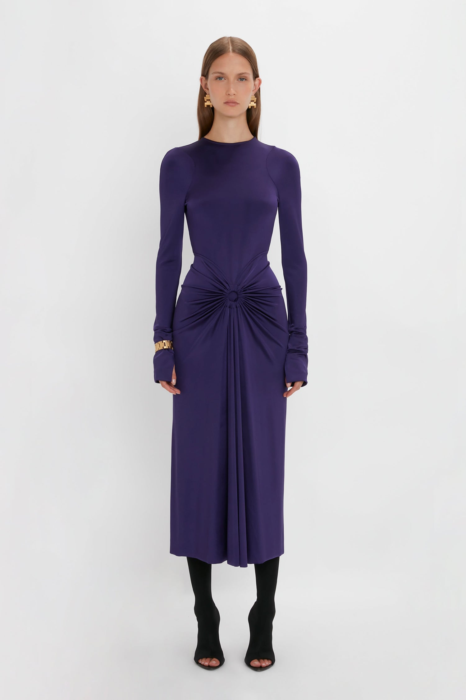 Woman in a long-sleeve, form-fitting purple Long Sleeve Gathered Midi Dress In Ultraviolet by Victoria Beckham made from body-sculpting stretch fabric stands against a plain white background, wearing black tights and open-toe heels, accessorized with gold earrings.