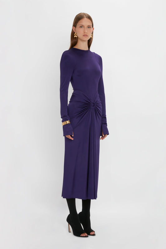 A person stands against a white background wearing a long-sleeved, purple Long Sleeve Gathered Midi Dress In Ultraviolet by Victoria Beckham with a front knot detail, black heeled sandals, gold bracelets, and small, round earrings. The sensuous and playful outfit is made from body-sculpting stretch fabric that enhances the silhouette.