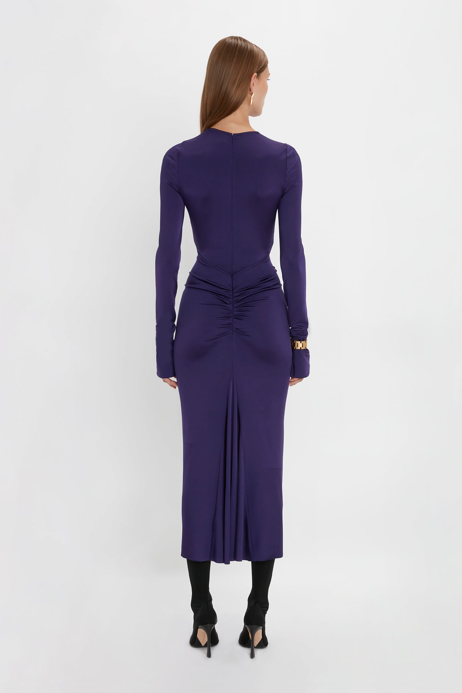 A woman with long brown hair, wearing the Long Sleeve Gathered Midi Dress In Ultraviolet by Victoria Beckham and black heels, is standing and facing away.