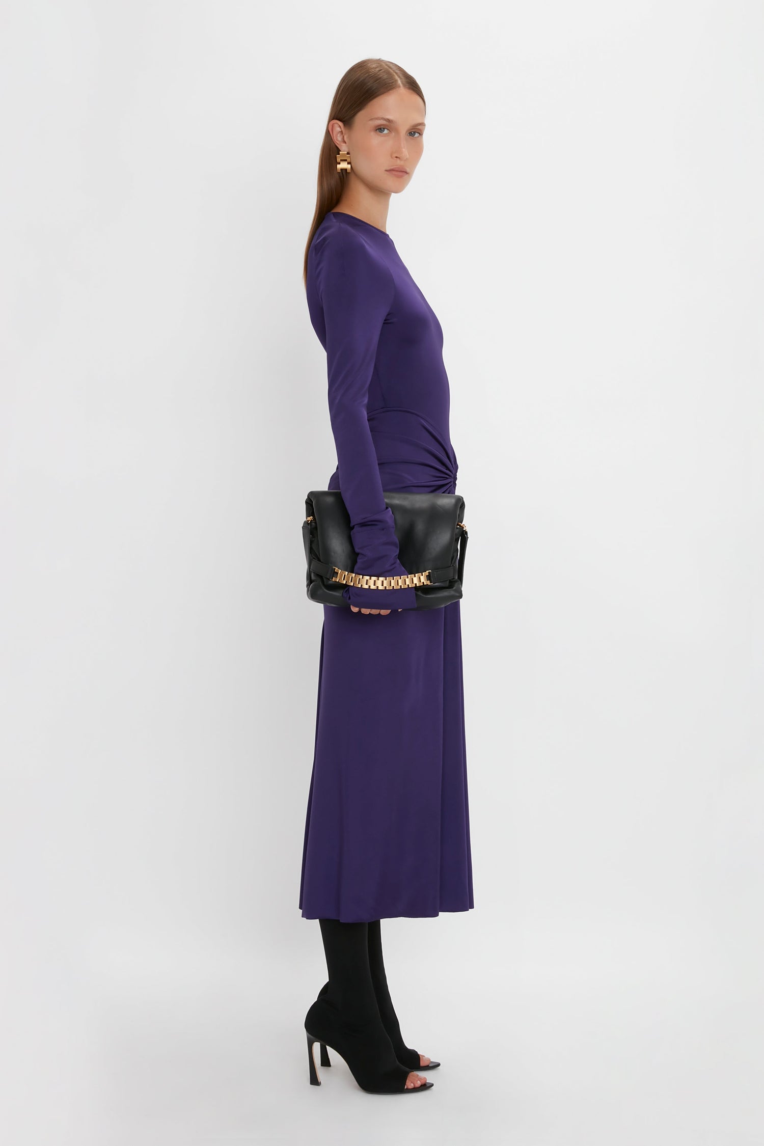 Woman standing in profile wearing a sensuous and playful, Victoria Beckham Long Sleeve Gathered Midi Dress In Ultraviolet, black tights, high-heeled open-toe shoes, and holding a black handbag with a gold chain. White background.