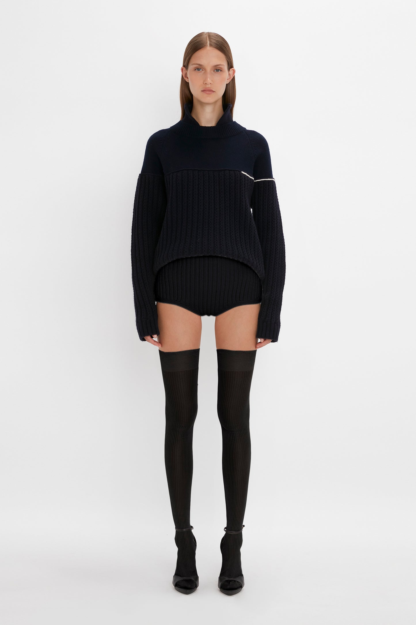 A woman in a black turtleneck sweater dress and Victoria Beckham Exclusive Over The Knee Socks In Black stands against a white background.