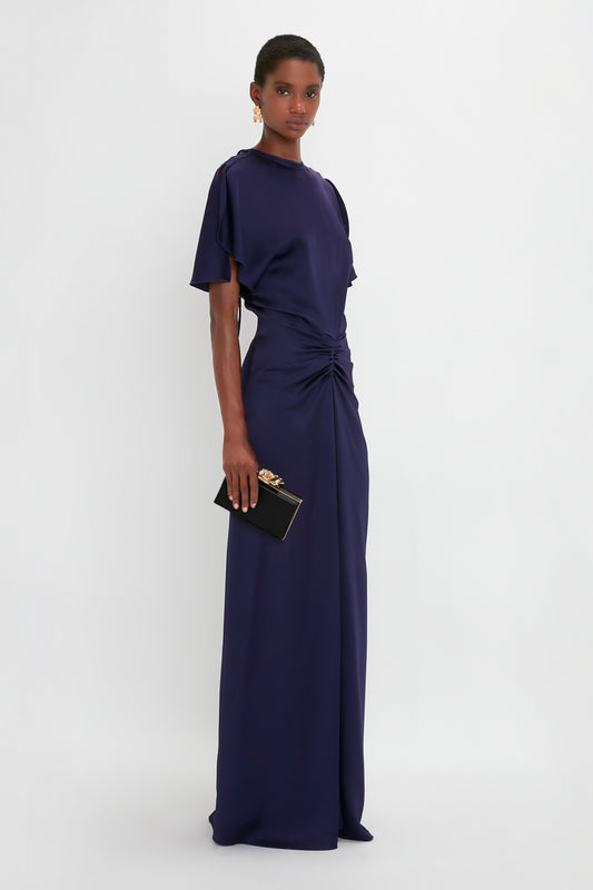 A woman stands against a white background, wearing a navy blue, floor-length gown with short sleeves and holding an exquisite black clutch featuring the Frame Flower Minaudiere Bag in Black by Victoria Beckham.