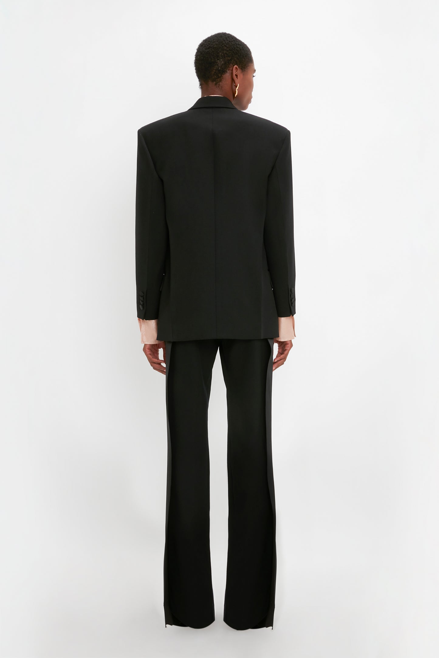 Person standing with their back to the camera, wearing an oversized black Satin Lapel Tuxedo Jacket in Black by Victoria Beckham and flare pants, against a white background.