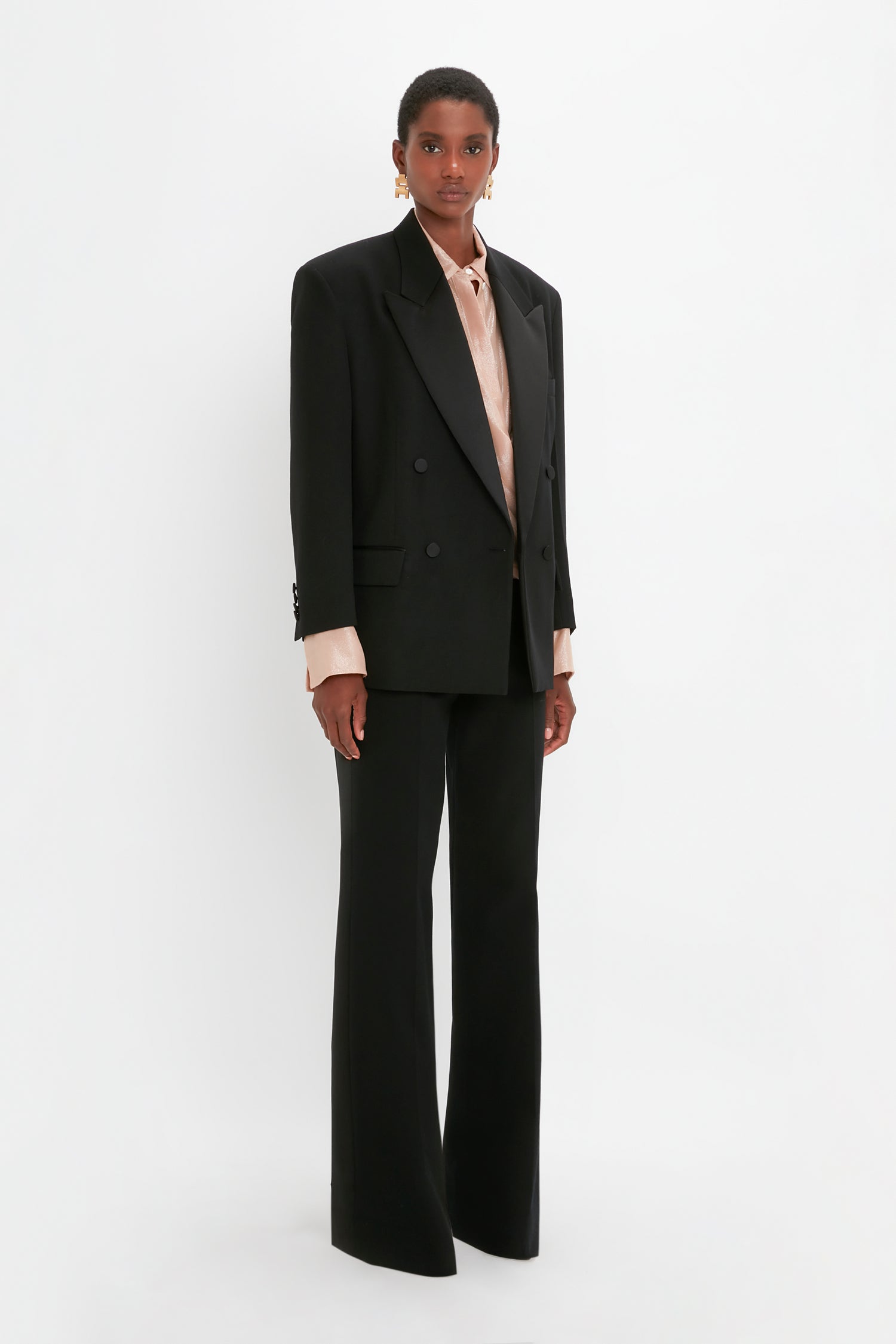 Person standing against a white background wearing an oversized black Satin Lapel Tuxedo Jacket in Black by Victoria Beckham, matching wide-leg trousers, satin gold earrings, and a light pink blouse.