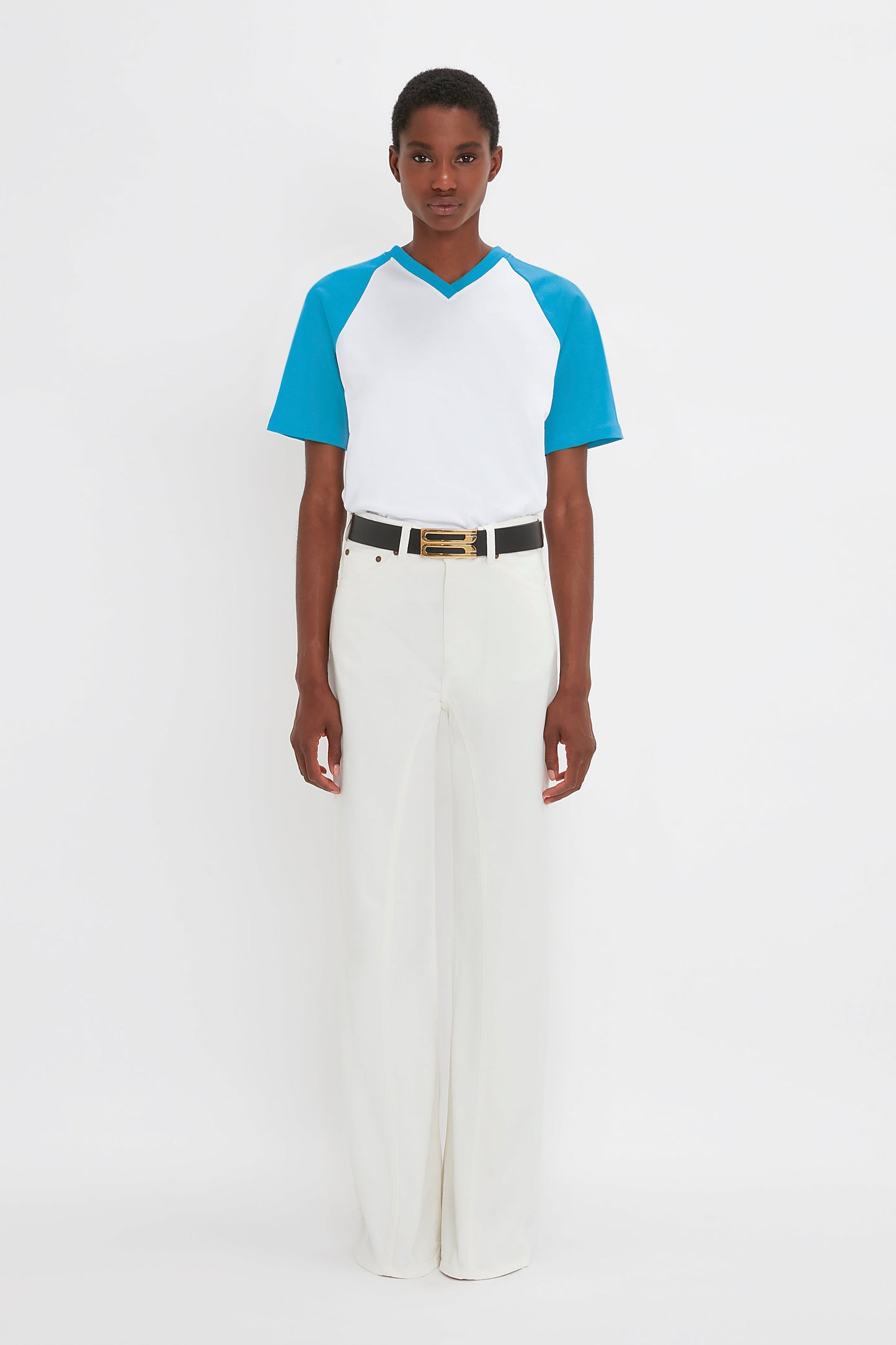 A person stands against a plain white background, donning an Exclusive Football T-Shirt in Blue by Victoria Beckham made from organic cotton, paired with white pants and accented by a black belt with a gold buckle.