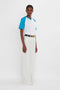 A person stands against a white background, wearing an Exclusive Football T-Shirt In Blue by Victoria Beckham, white wide-leg trousers, and a black belt.