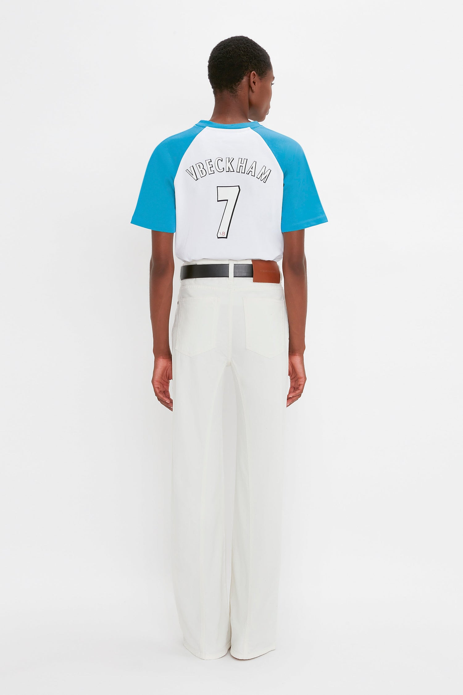 Person facing away, wearing a blue and white **Victoria Beckham Exclusive Football T-Shirt In Blue** with "V. BECKHAM 7" on the back, made from organic cotton, paired with white pants.