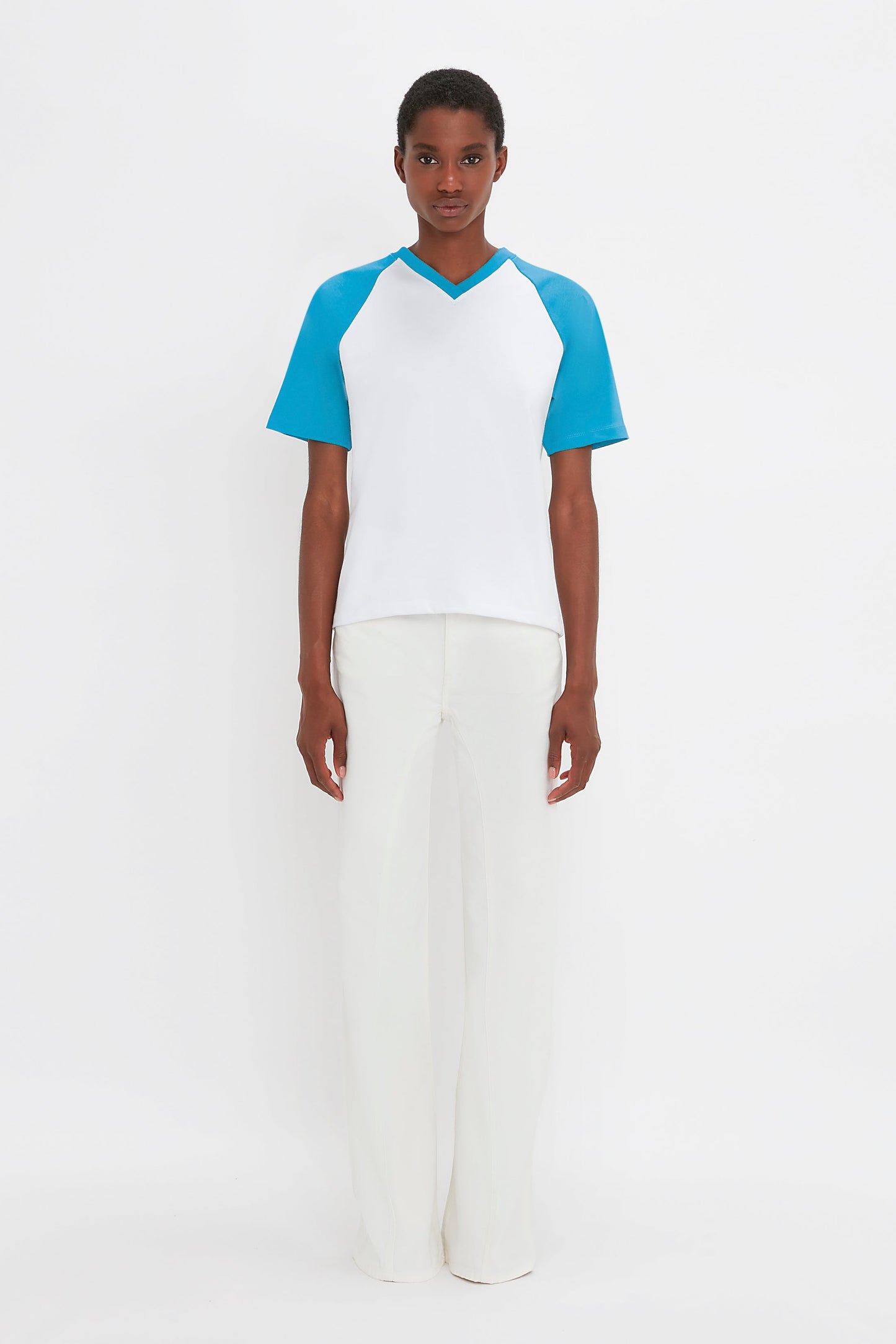 A person standing against a white background, wearing the Victoria Beckham Exclusive Football T-Shirt In Blue made of organic cotton, paired with white pants.