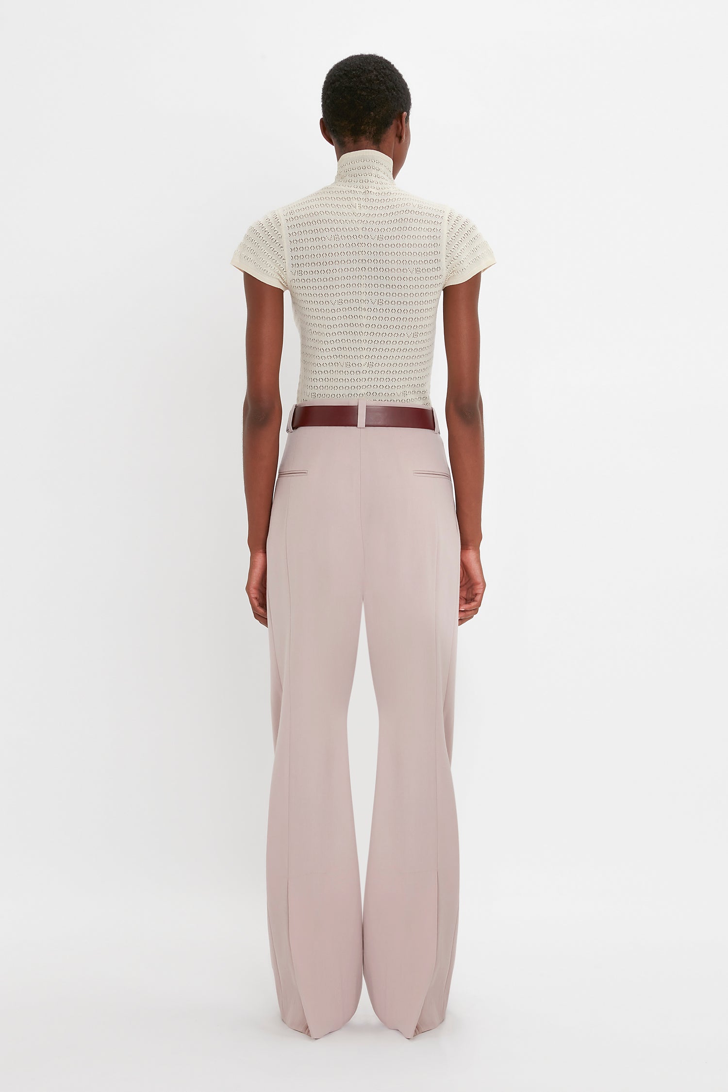 A person is standing with their back to the camera, wearing a beige Polo Neck Knitted T-Shirt In Cream by Victoria Beckham and light pink wide-leg pants with a brown belt against a plain white background. The outfit features trans-seasonal appeal, perfect for mixing and matching throughout the year.