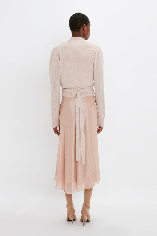 A person is standing with their back to the camera, wearing a light pink, Victoria Beckham Wrap Front Cardigan In Nougat, a matching high-waisted skirt, and beige heels against a plain white background.