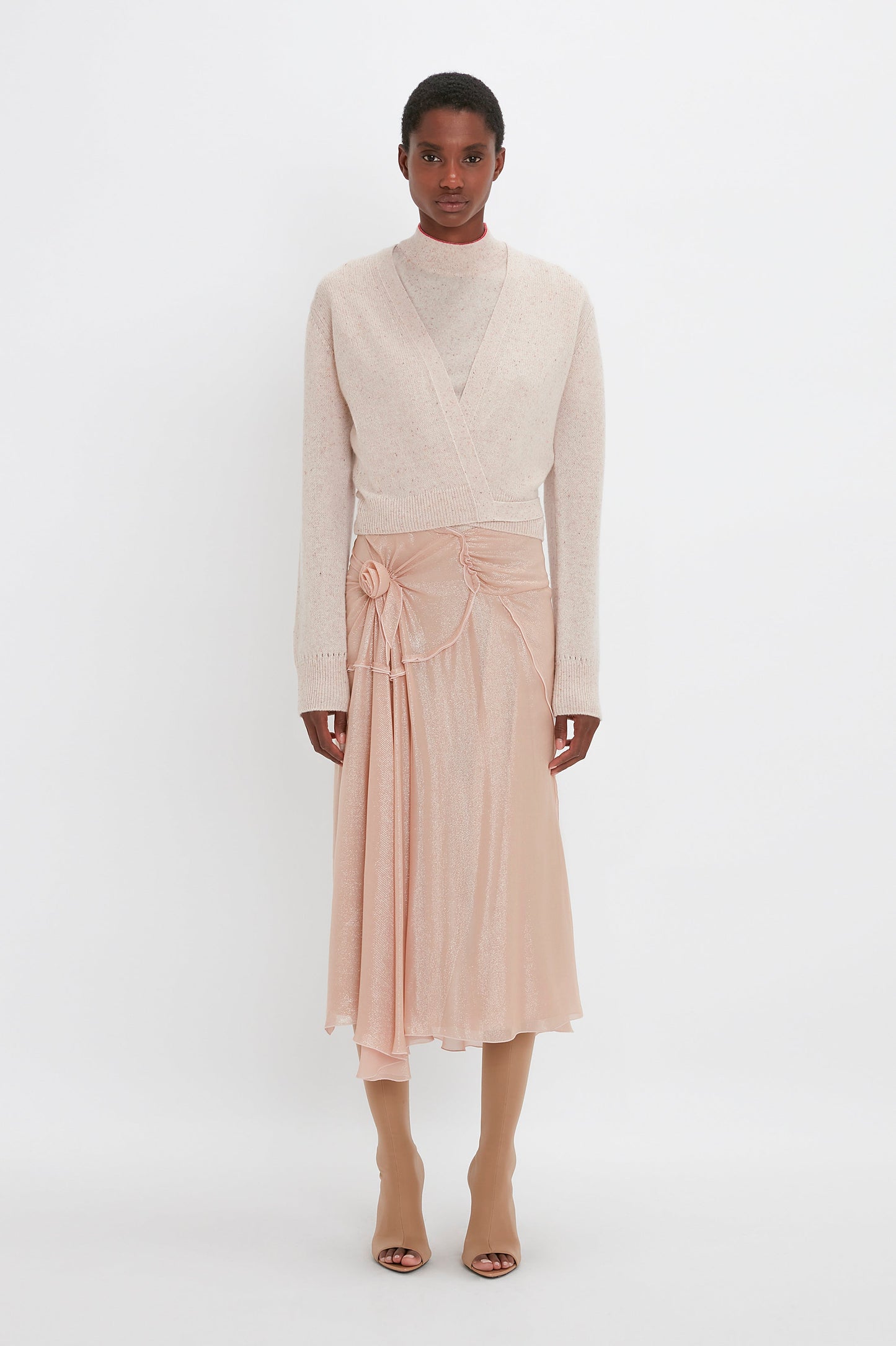 A person stands against a plain white background wearing a light-colored wrap sweater, a high-neck top, a Flower Detail Cami Skirt In Rosewater by Victoria Beckham, and beige boots.