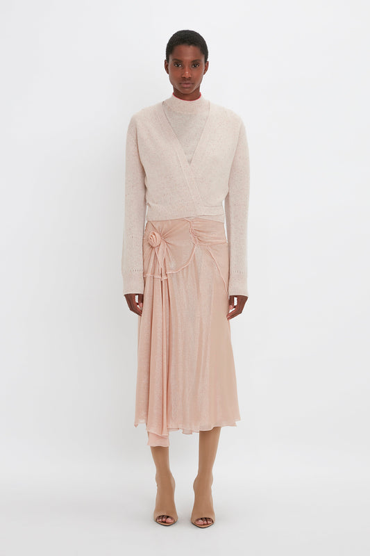 Person standing against a plain white background, wearing a Victoria Beckham-inspired light beige lightweight **Victoria Beckham Wrap Front Cardigan In Nougat** over a turtleneck and a light pink skirt with a draped design, paired with beige tights and sandals.