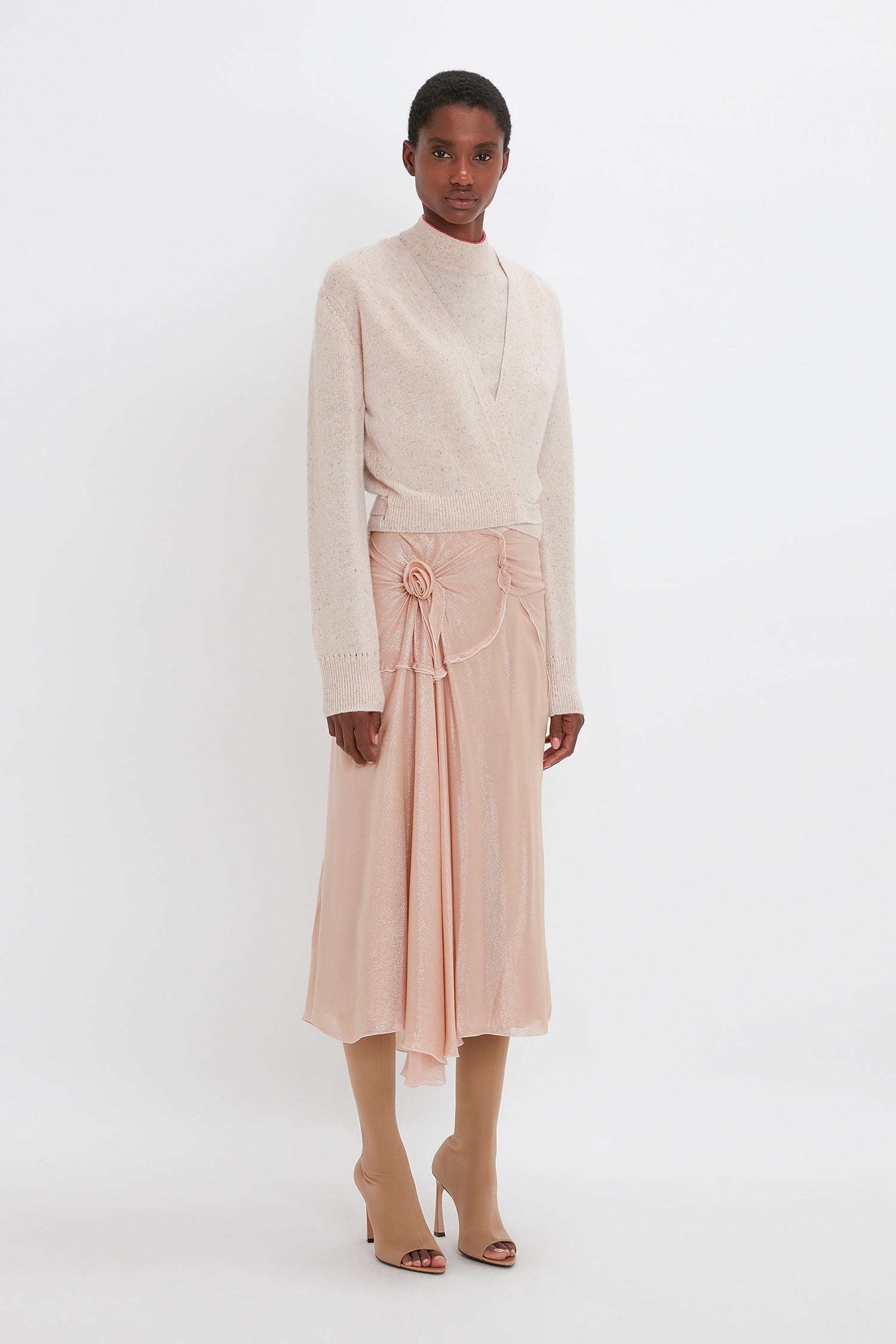 A person stands against a white background, wearing a cream sweater, a light pink Flower Detail Cami Skirt In Rosewater by Victoria Beckham, and tan open-toe boots.