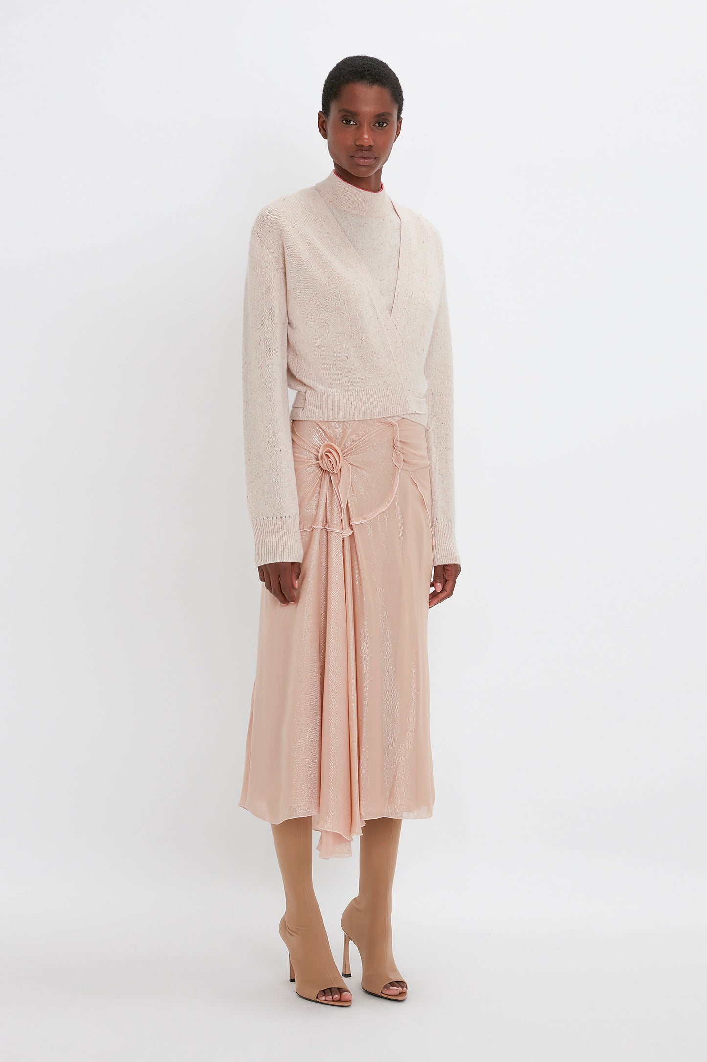 A person stands against a plain backdrop wearing a Victoria Beckham Wrap Front Cardigan In Nougat, a flowing peach midi skirt with floral detail, and beige heeled boots.