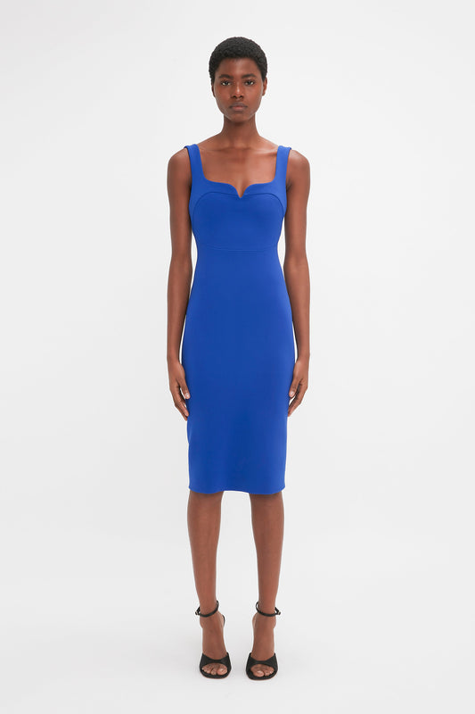 A person stands against a plain background, wearing a form-fitting Sleeveless Fitted T-Shirt Dress In Palace Blue by Victoria Beckham and black high-heeled sandals.