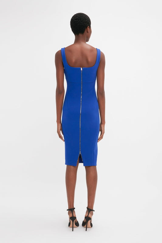 Person standing facing away, wearing a Victoria Beckham Sleeveless Fitted T-Shirt Dress in Palace Blue with a gold zipper down the back and black high-heeled shoes.