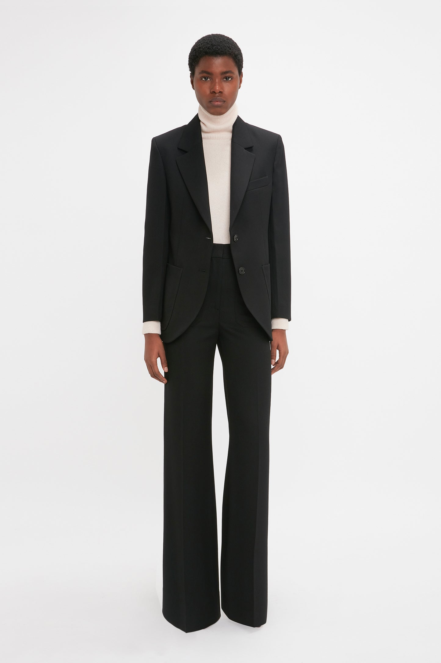 Person wearing a Victoria Beckham Patch Pocket Jacket In Black, white turtleneck, and black wide-leg trousers stands against a white background.