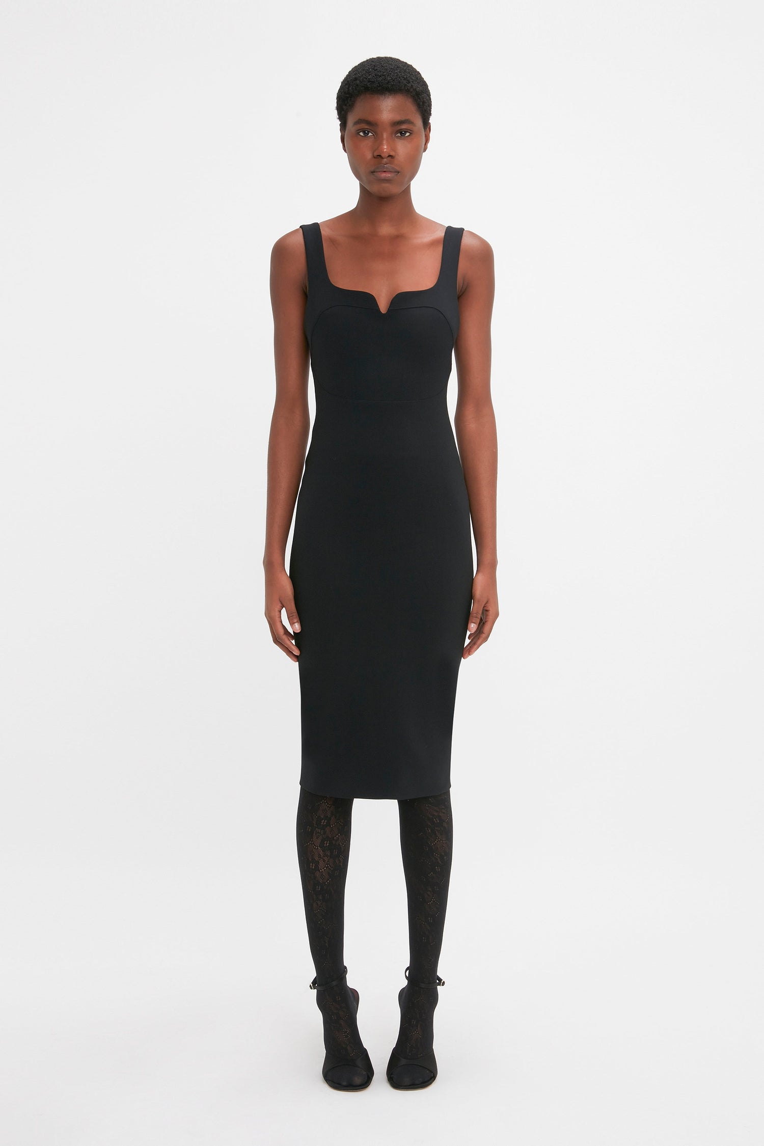 A person stands facing forward, wearing a Sleeveless Fitted T-Shirt Dress In Black by Victoria Beckham, featuring an exposed double-ended zip and black patterned stockings, set against a plain white background.