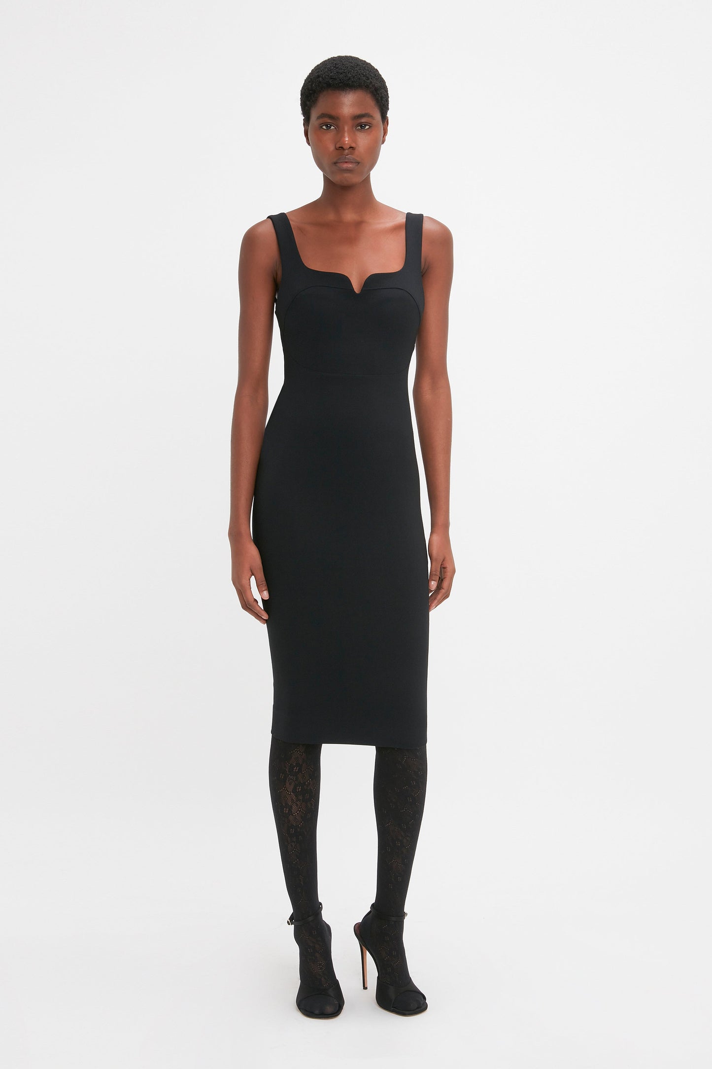 A person stands against a plain white background, wearing a form-fitting black **Sleeveless Fitted T-Shirt Dress In Black by Victoria Beckham**, complemented by black patterned stockings and black high-heeled shoes.