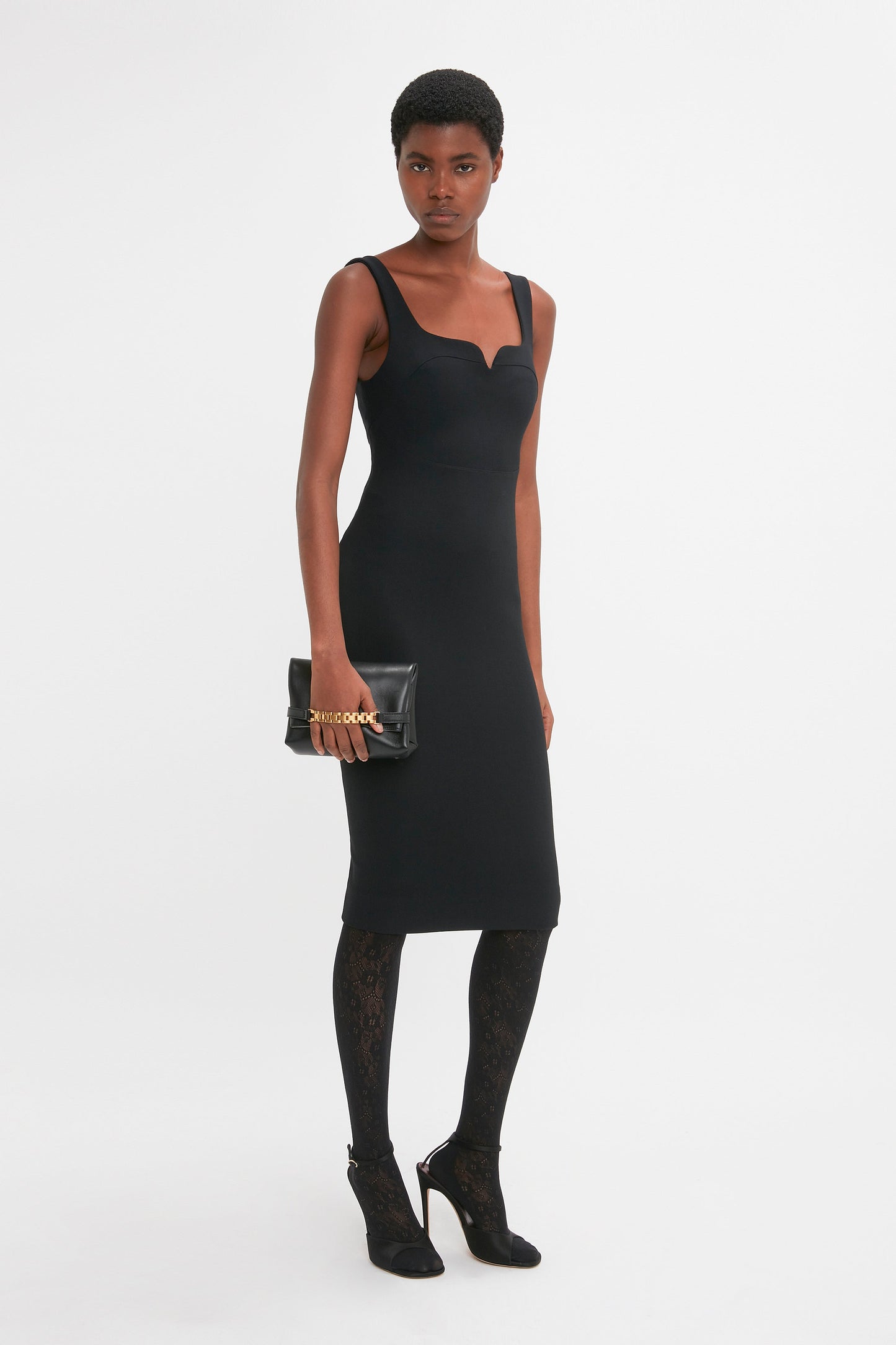 A person wearing a Victoria Beckham Sleeveless Fitted T-Shirt Dress In Black, patterned black tights, and black heeled shoes holds a black clutch purse.