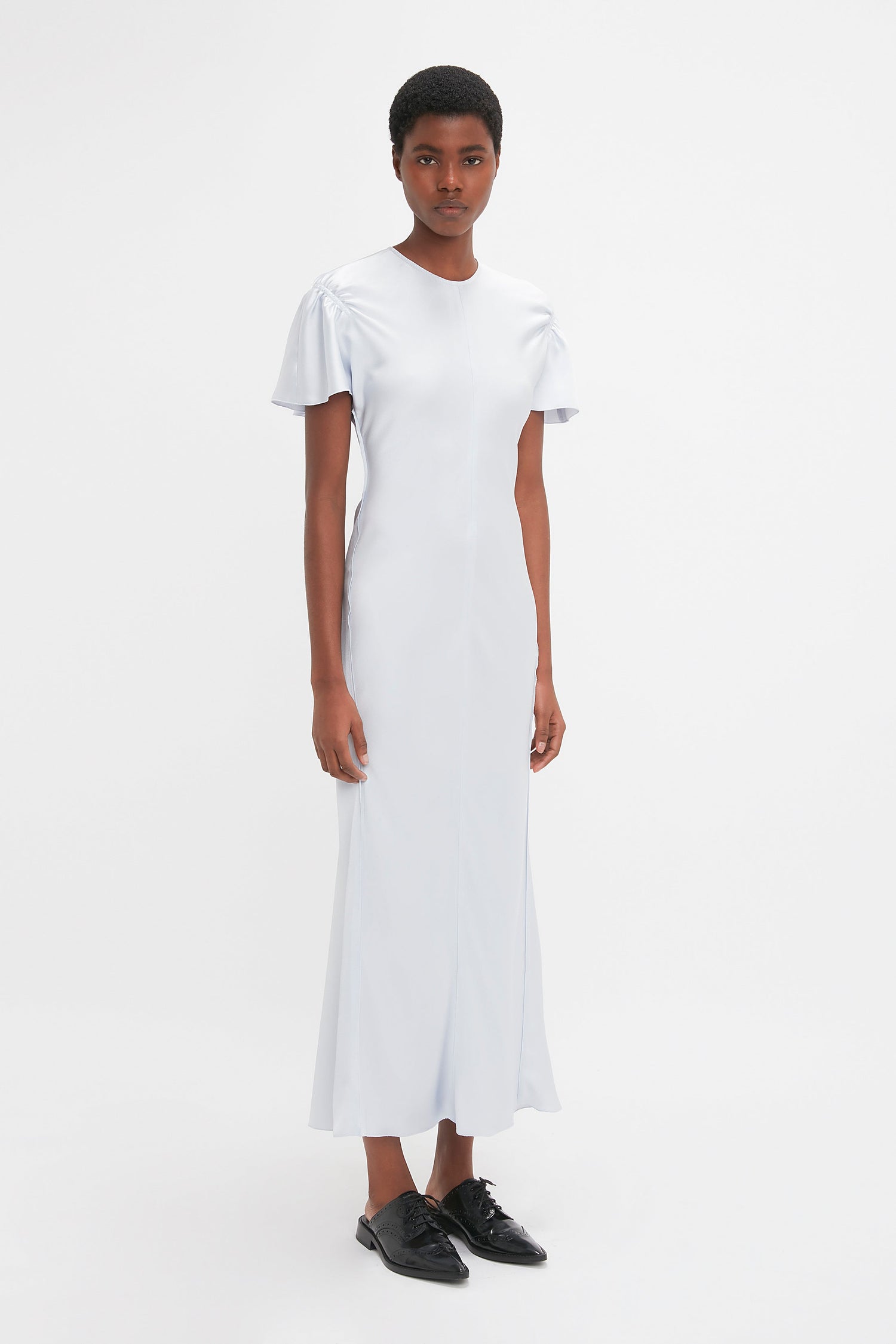 A person with short hair is standing against a plain white background, wearing an elegant Gathered Sleeve Midi Dress In Ice by Victoria Beckham and black shoes.