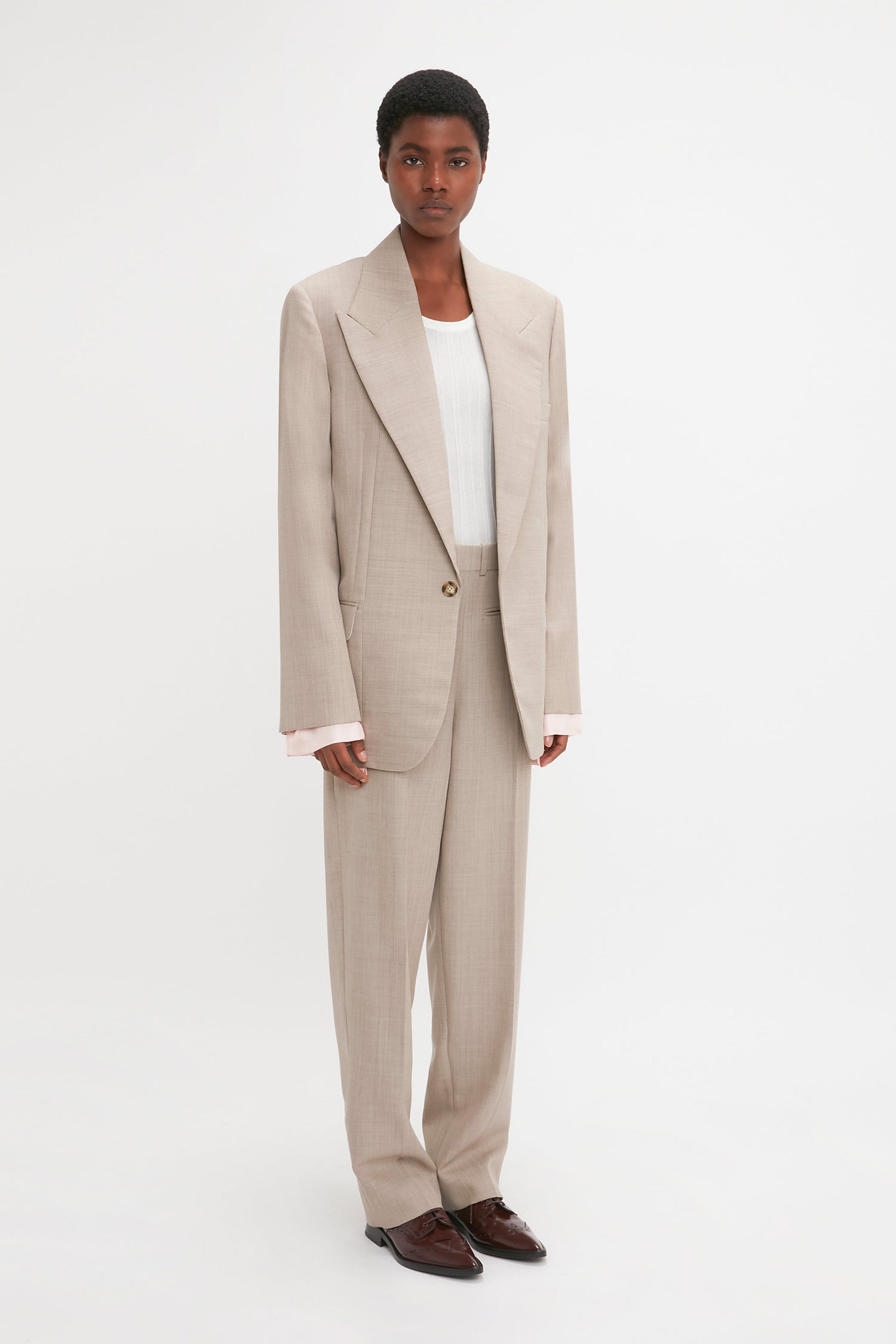 A person wearing a beige pure wool suit with a Darted Sleeve Tailored Jacket In Sesame by Victoria Beckham, complemented by a white shirt and brown shoes, stands against a white background.