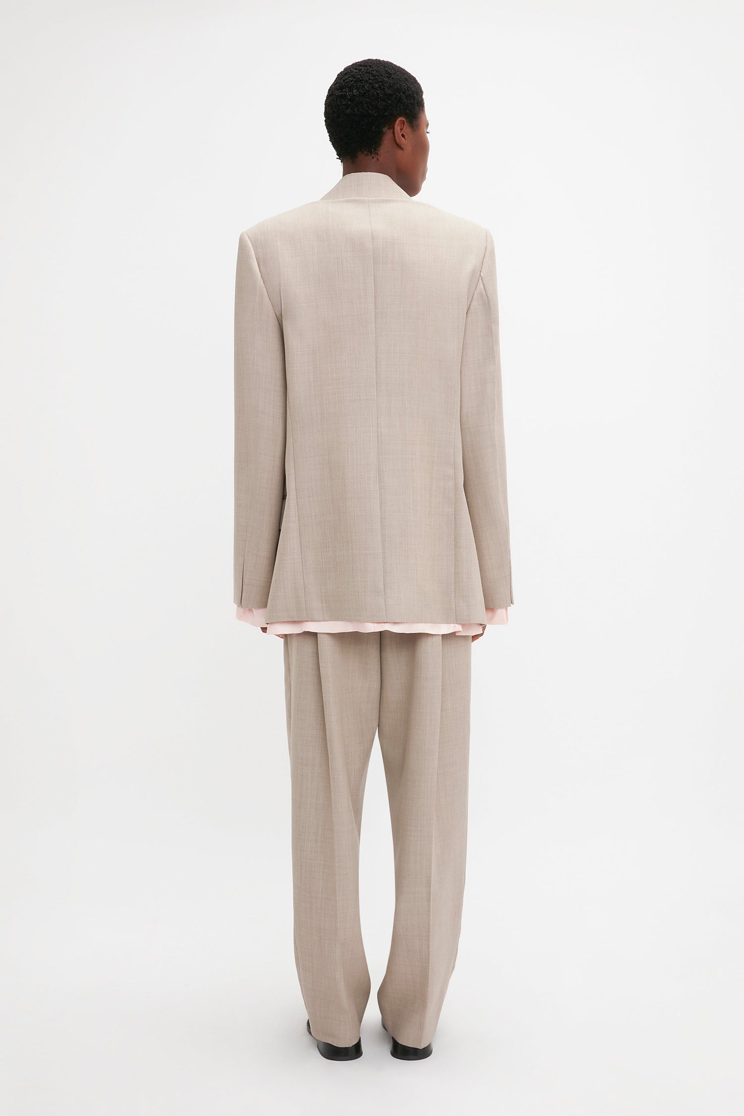 A person stands facing away from the camera wearing a beige Victoria Beckham Darted Sleeve Tailored Jacket In Sesame with contemporary detailing and black shoes, against a plain white background.