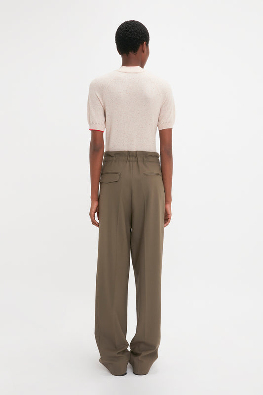 Person with short curly hair wearing a Short Sleeve Top In Nougat by Victoria Beckham and high-waisted, loose-fitting brown pants, featuring a delicate pointelle lace stitch pattern, standing with their back facing the camera against a plain white background.