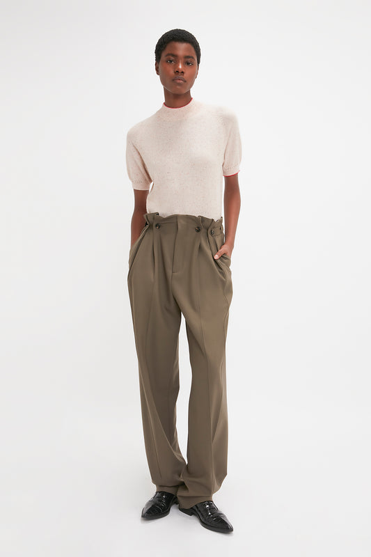 A person with short hair stands with hands in the pockets of high-waisted, olive-green trousers, wearing a Short Sleeve Top In Nougat by Victoria Beckham and black shoes. The background is plain white.