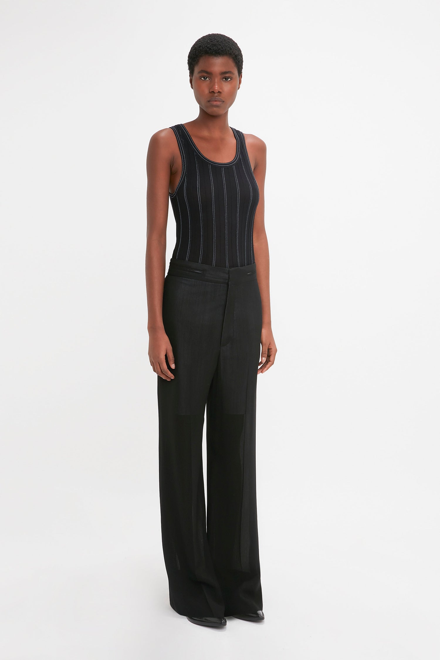 Person standing against a white background, wearing a Fine Knit Vertical Stripe Tank In Black-Blue by Victoria Beckham and black wide-leg pants, with a neutral expression.