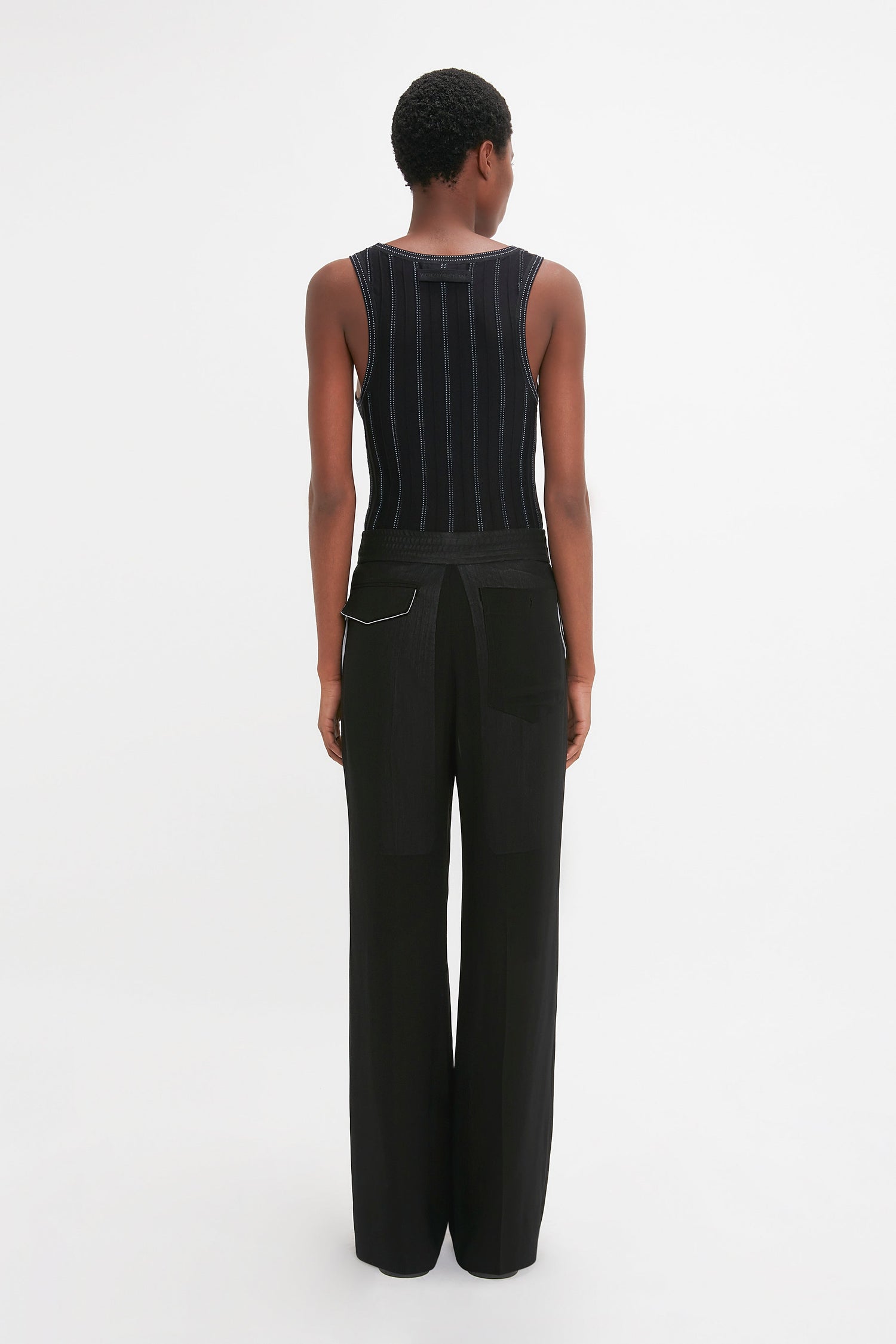 Person standing facing away from the camera, wearing a Fine Knit Vertical Stripe Tank In Black-Blue and black pants against a plain white background, resembling the sleek elegance of Victoria Beckham’s style.