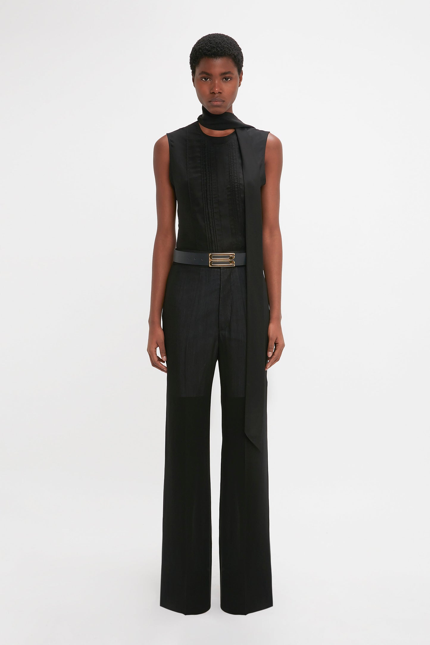 A person stands against a plain white background, wearing a Victoria Beckham Sleeveless Tie Neck Top In Black and black high-waisted, wide-leg pants.