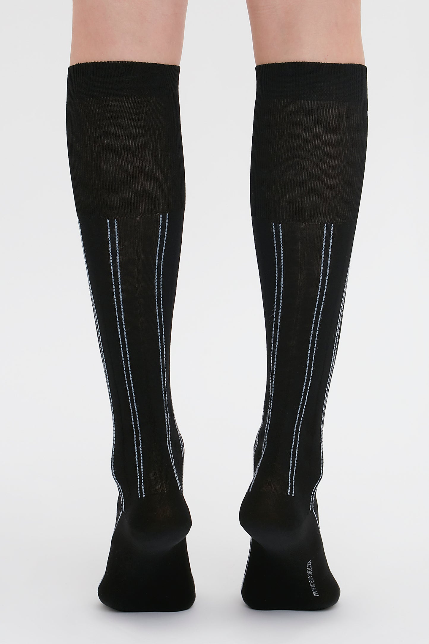 Back view of a person wearing Victoria Beckham Superfine Rib Socks In Black, knee-high 100% cotton socks with vertical steel blue and white stripes on the back.