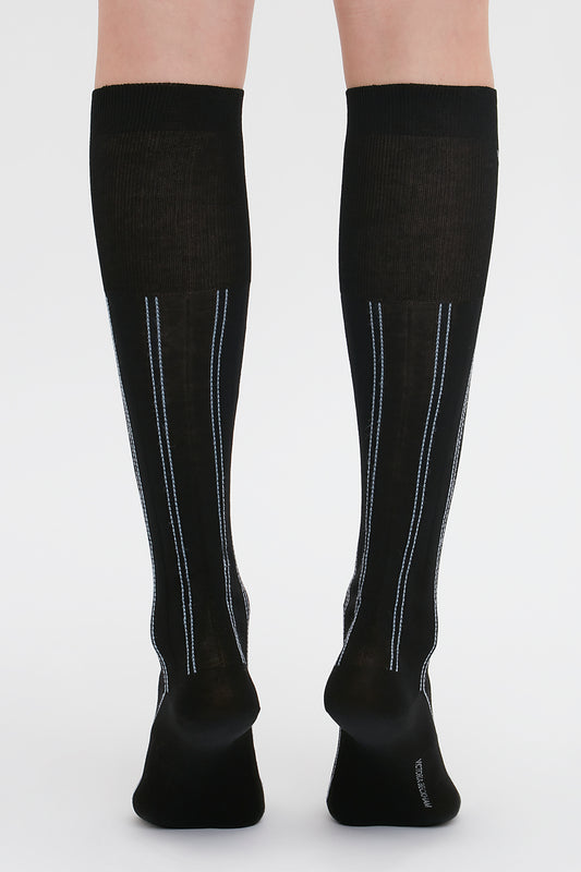 Back view of a person wearing Victoria Beckham Superfine Rib Socks In Black, knee-high 100% cotton socks with vertical steel blue and white stripes on the back.