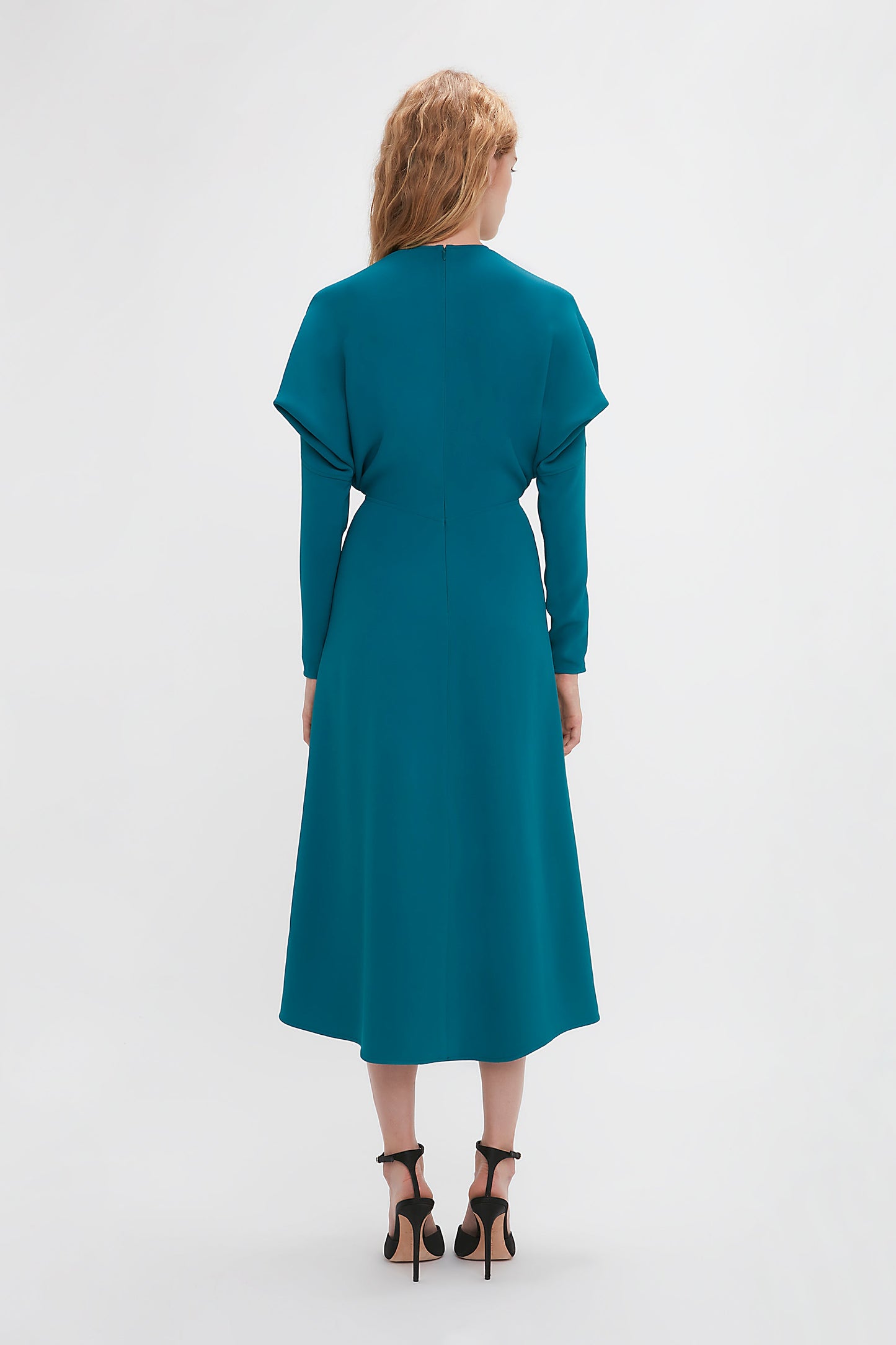 A person with long blonde hair is seen from the back, elegantly wearing a Victoria Beckham Long Sleeve Dolman Midi Dress In Petroleum made of luxurious cady fabric and black high-heeled sandals, exuding contemporary sophistication against a plain white background.