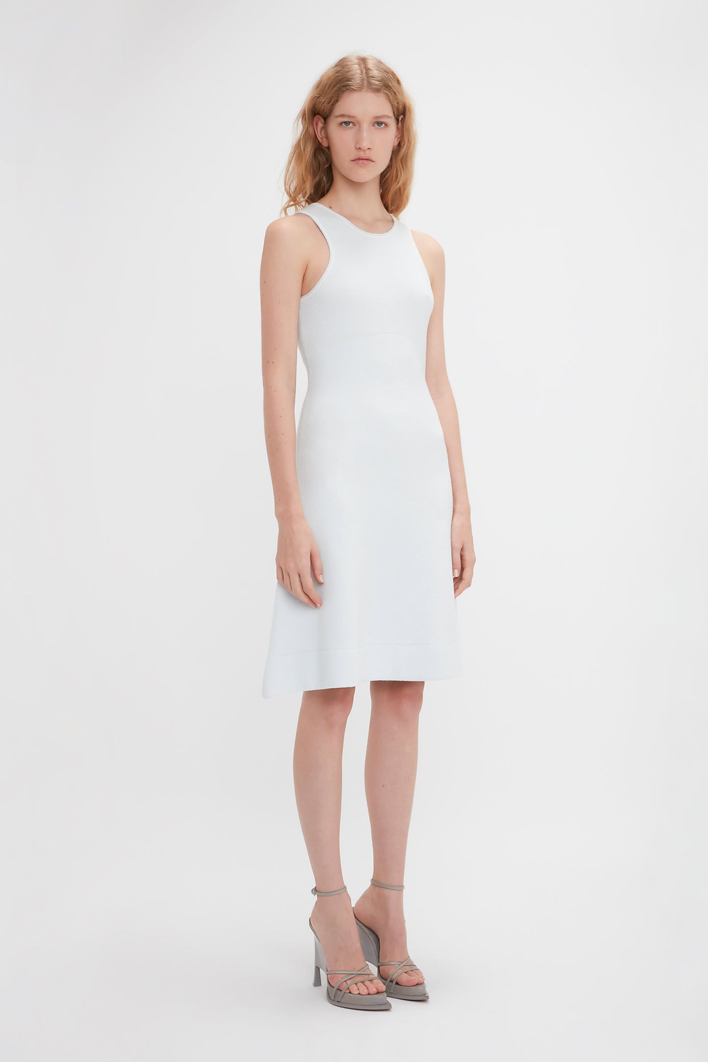 A woman with long blonde hair is standing and wearing a Sleeveless Tank Dress In Ice by Victoria Beckham paired with high-heeled sandals. She is looking directly at the camera against a plain white background.