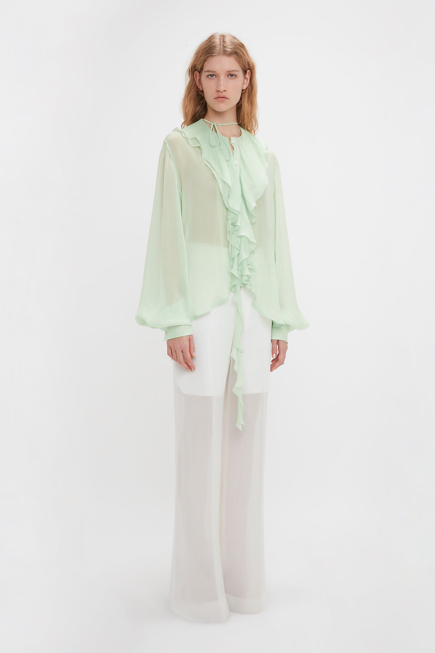 Young woman modeling the Victoria Beckham Romantic Blouse In Jade, a sheer light green hue with blouson sleeves and ruffles, paired with flowing white trousers against a plain white background.