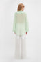 A person with light-colored hair is seen from the back, wearing a semi-sheer, light green Romantic Blouse In Jade by Victoria Beckham with blouson sleeves and white wide-leg pants against a plain white background.