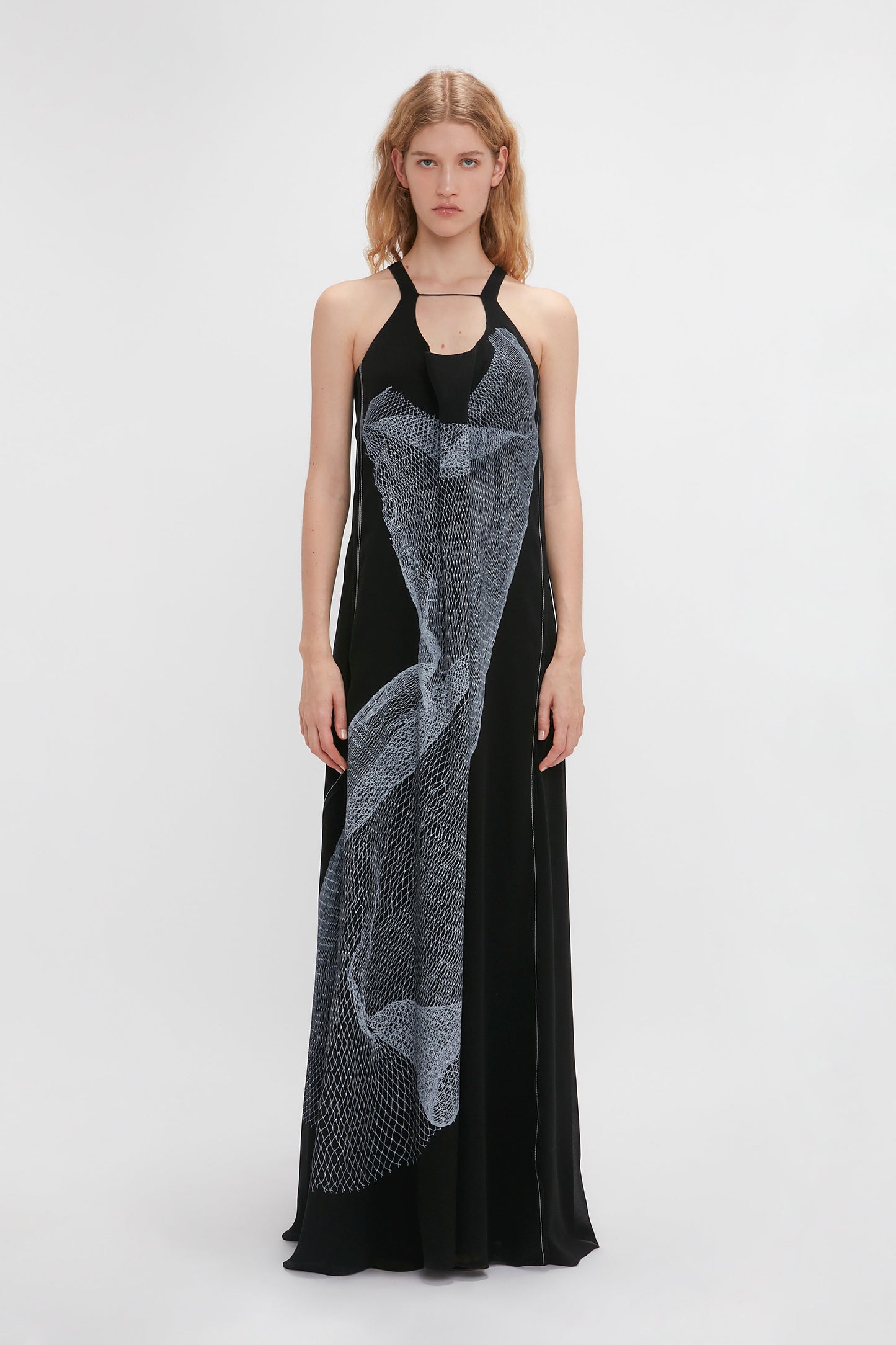 A person stands against a white background wearing the Sheer Cami Gown In Black-White Contorted Net by Victoria Beckham, featuring a contemporary twisted net print draped diagonally across the front, echoing the SS24 runway highlight.