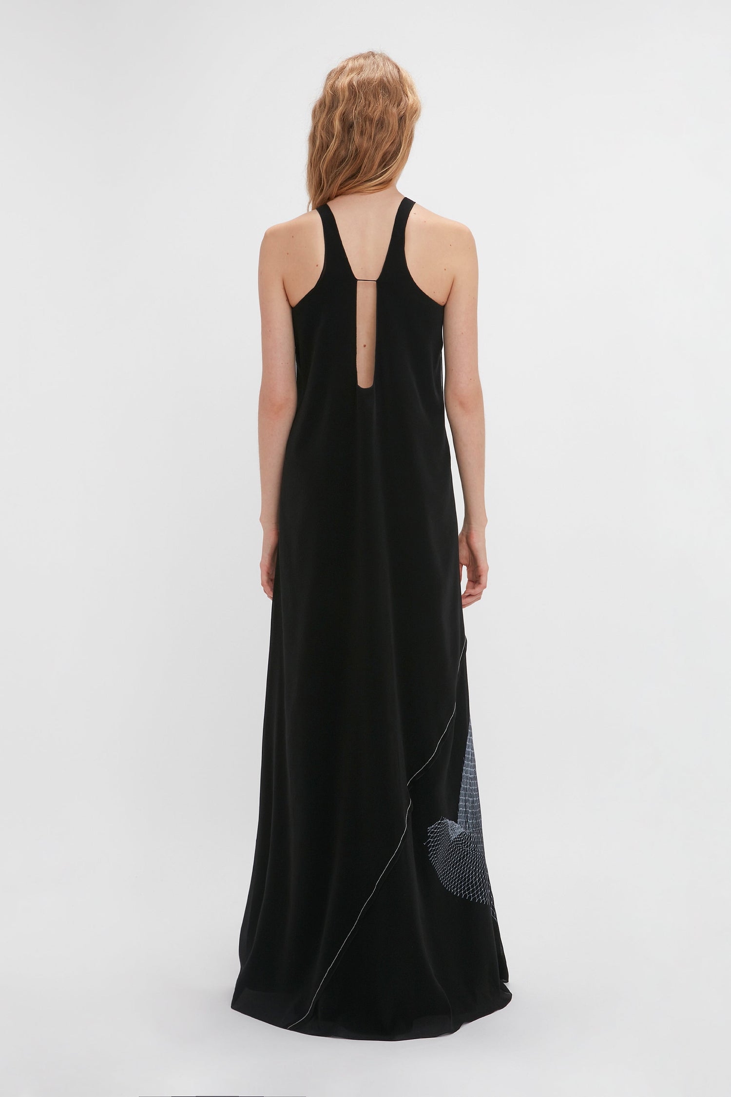 A woman with wavy hair wearing a long, black sleeveless dress is photographed from the back against a plain white background, showcasing a Victoria Beckham Sheer Cami Gown In Black-White Contorted Net that is set to be an SS24 runway highlight.