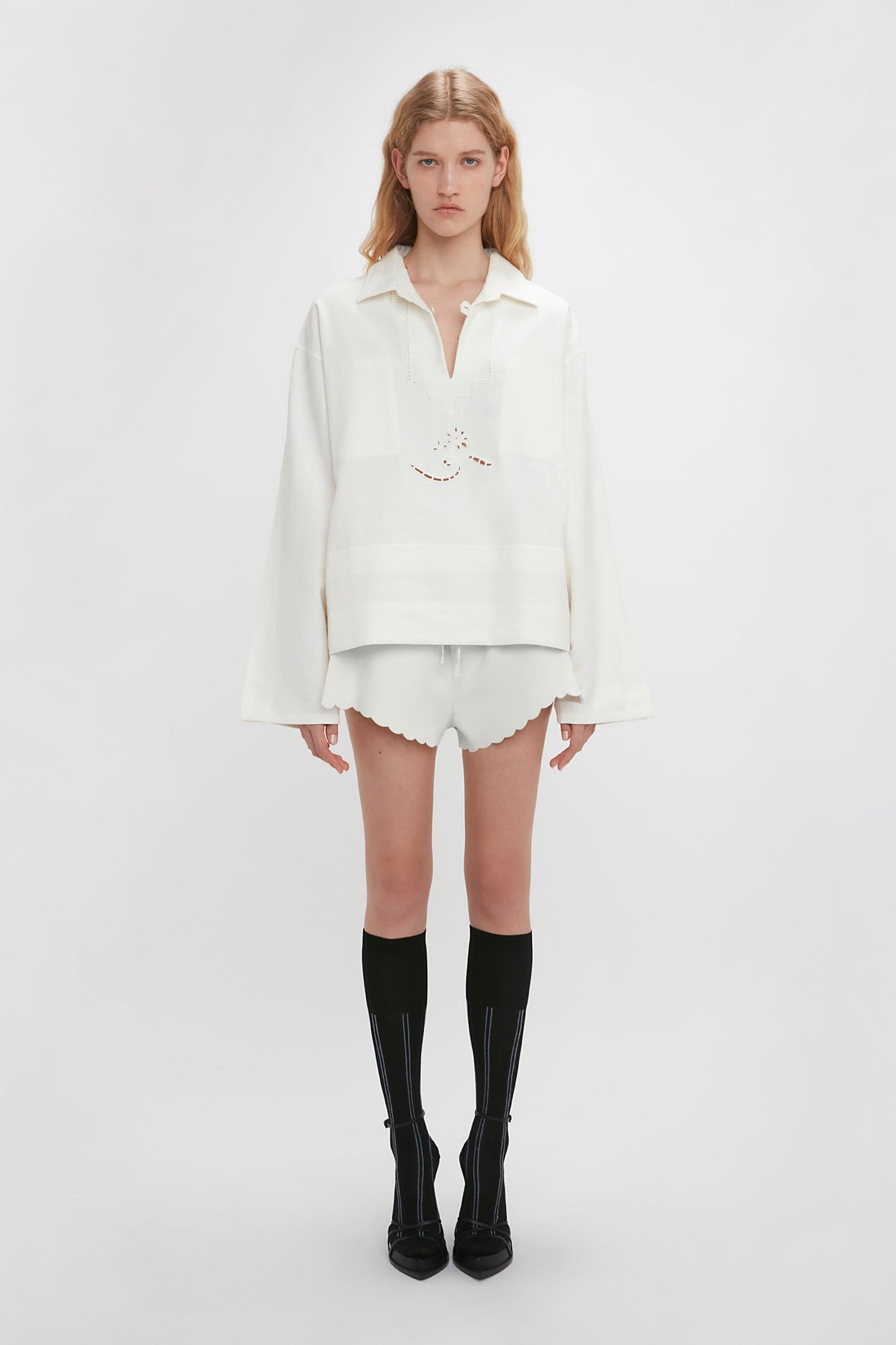 A person stands against a plain background wearing a white blouse with delicate embroidered detailing, Drawstring Embroidered Mini Short In Antique White by Victoria Beckham, and knee-high black socks with black shoes.