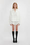 A person stands against a plain background wearing a white blouse with delicate embroidered detailing, Drawstring Embroidered Mini Short In Antique White by Victoria Beckham, and knee-high black socks with black shoes.