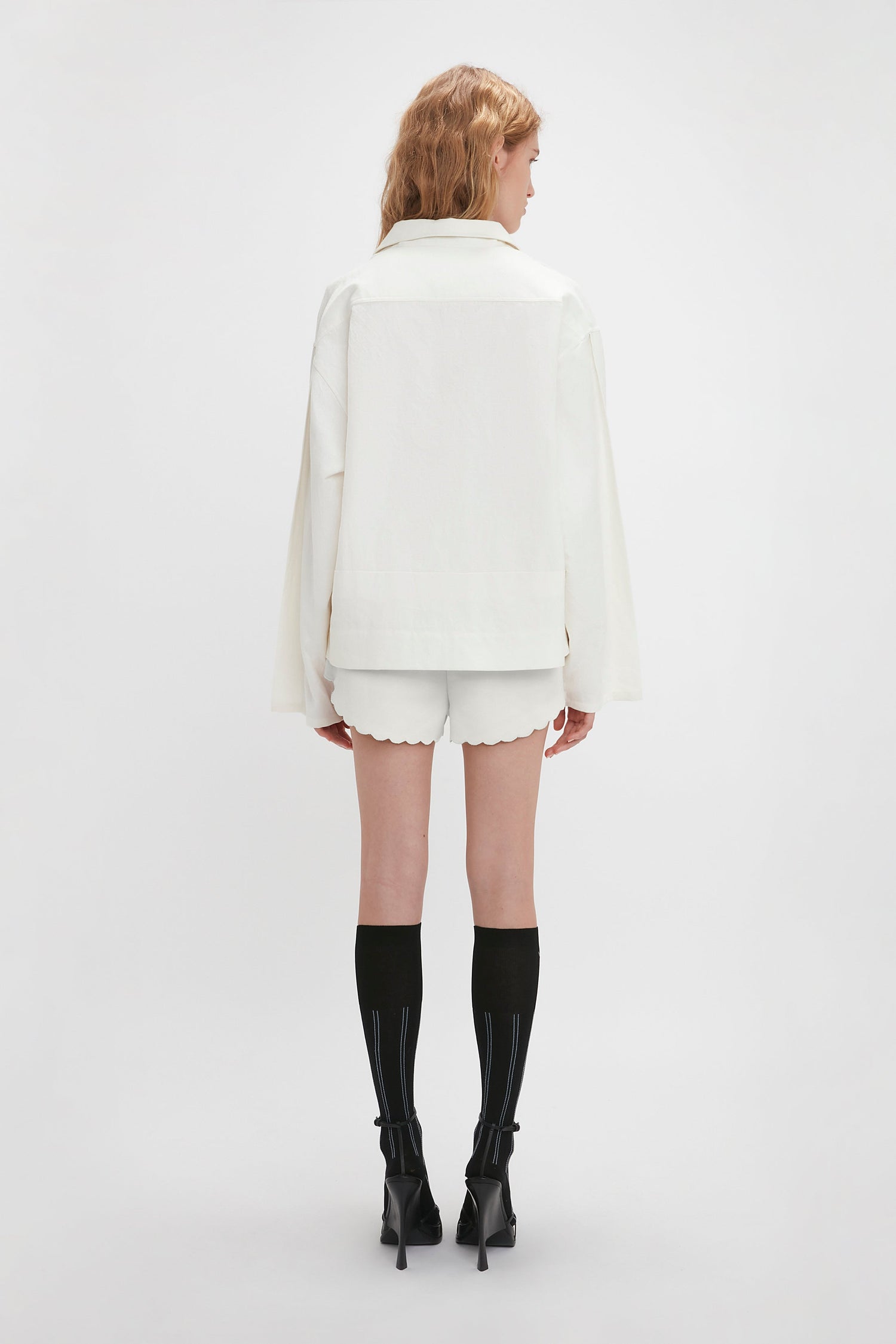 Rear view of a person with blonde hair wearing a white jacket with embroidered detailing, Drawstring Embroidered Mini Short In Antique White by Victoria Beckham, black knee-high socks, and black high-heeled shoes, standing against a plain white background.