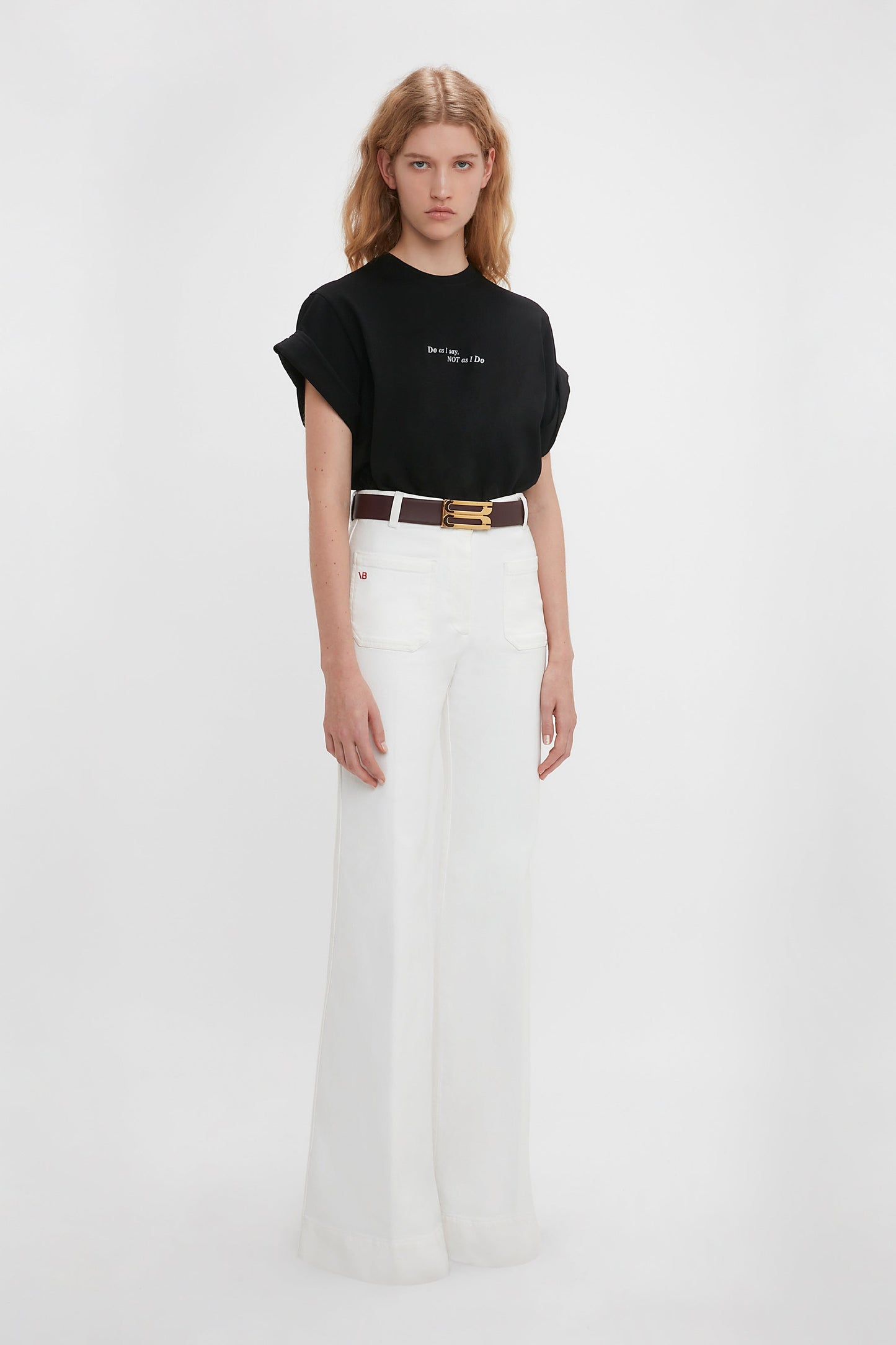 Young woman in a Victoria Beckham 'Do As I Say, Not As I Do' Slogan T-shirt and white high-waisted flared jeans, standing against a plain white background.