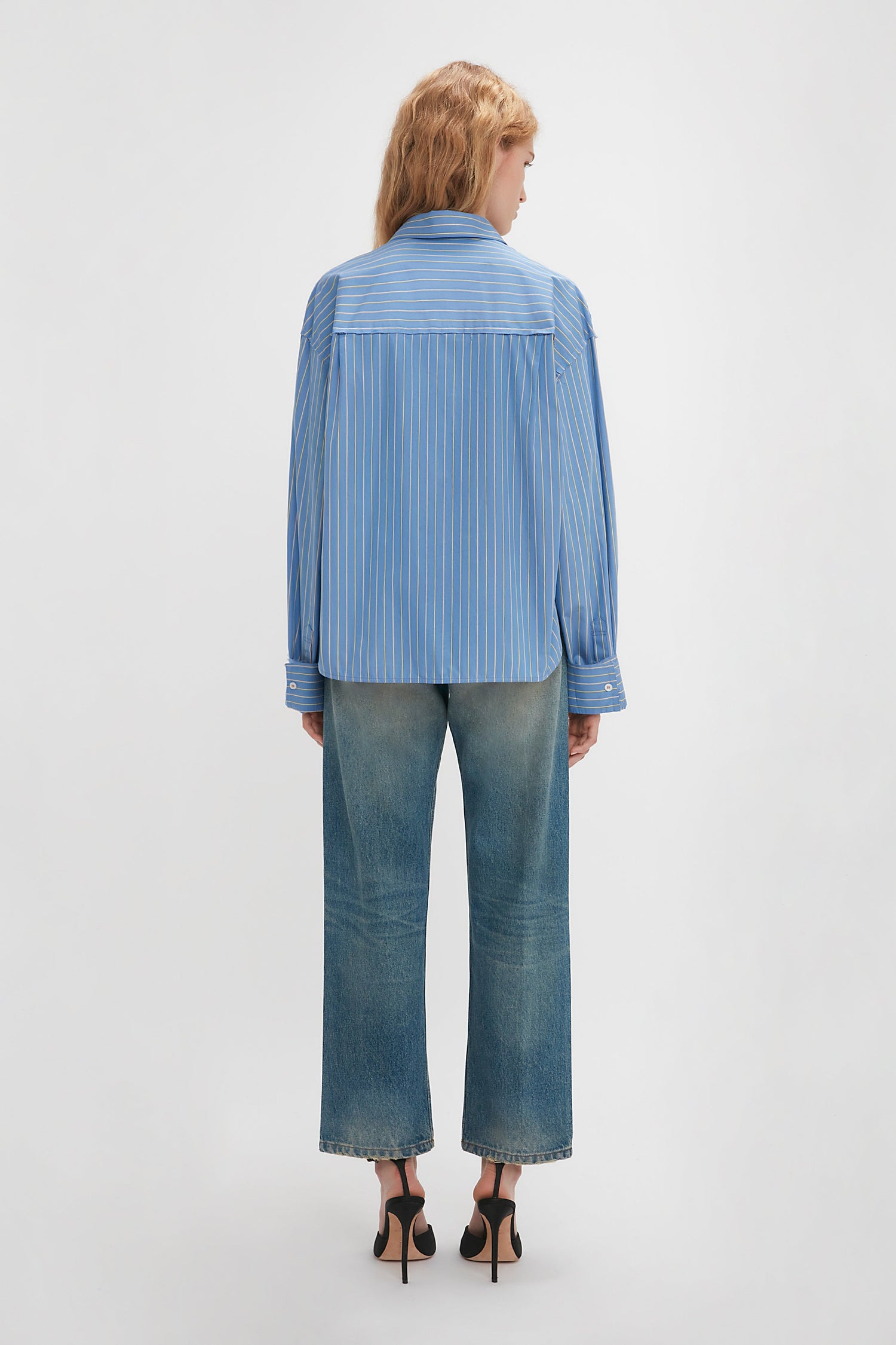 Person with long hair in a Cropped Seam Detail Shirt In Steel Blue by Victoria Beckham and faded jeans, standing with their back to the camera, exuding a utilitarian sensibility.