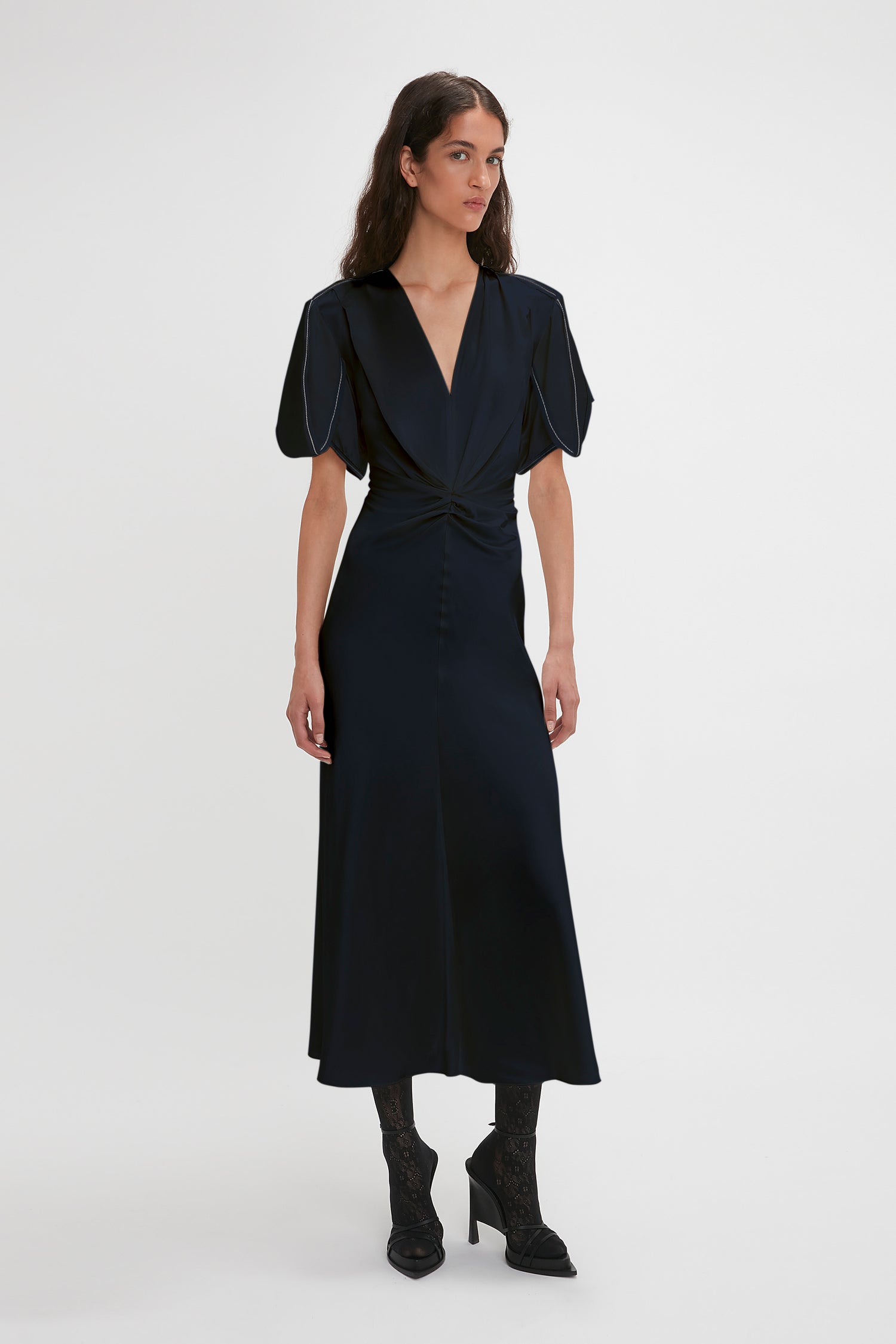 A woman stands against a white background wearing an Exclusive Gathered V-Neck Midi Dress In Navy by Victoria Beckham, with puff sleeves, paired with black lace-up boots.