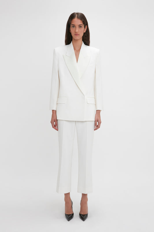 A woman is standing against a plain background, wearing a white blazer and matching trousers with black V Pump In Black Brushed Leather by Victoria Beckham featuring a sculptural heel. She has long, dark hair and a straight posture.