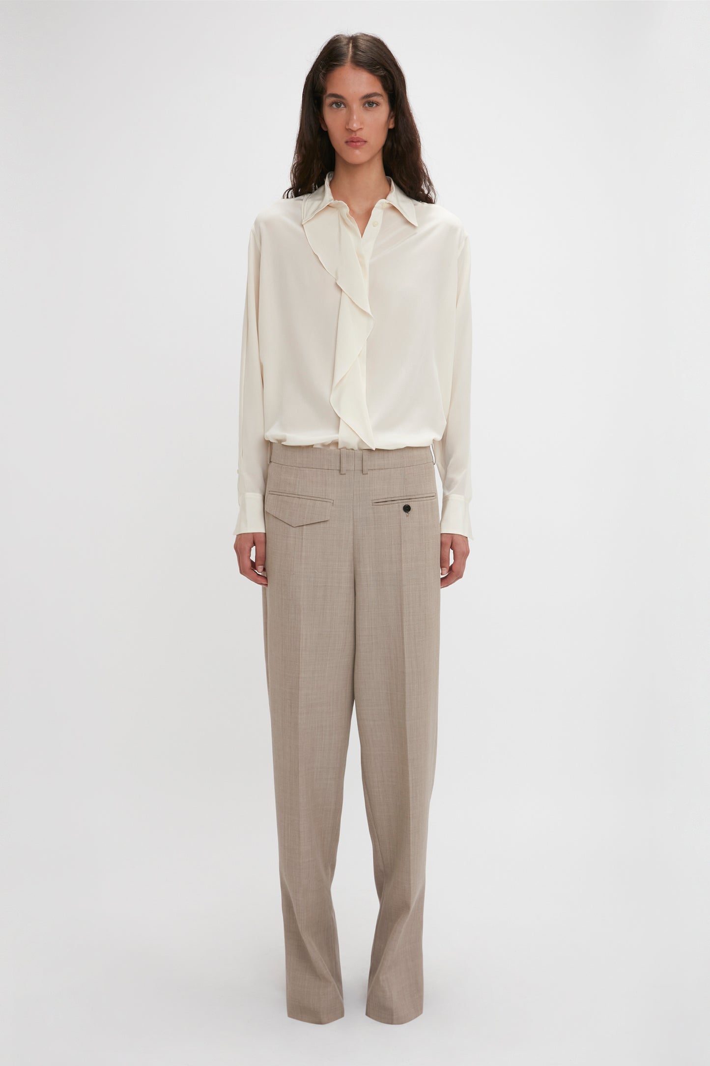 A person stands against a plain background, wearing an Asymmetric Ruffle Blouse In Ivory by Victoria Beckham with ruffle detail and voluminous sleeves, paired with beige wide-leg trousers.